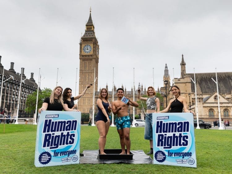 LUSH and EachOther activists stage a shower protest in parliament square. Two activists stand having a shower in their swimming costume and trunks, while others hold a shower head over them and big box mock ups of LUSH's 'human rights' product that reads: "human rights for everyone"
